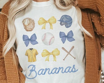 May include: White t-shirt featuring baseball-themed illustrations, including a baseball, glove, helmet, bows, and bats. The word "Bananas" is written in blue script. The shirt is a casual, sporty design.