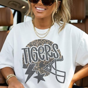 May include: White short-sleeve t-shirt with the word "TIGERS" in a distressed font, a leopard print football helmet graphic, a lightning bolt, and a star. The shirt is a casual, sporty style.