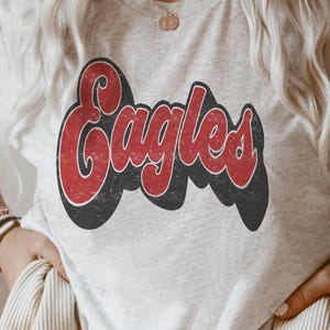 May include: A heather gray short-sleeve t-shirt featuring the word Eagles printed in a distressed red font with a thick black drop shadow. The graphic is centered on the chest of the shirt, which has a crew neckline and a soft, speckled texture.
