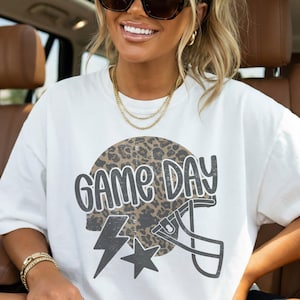 May include: White t-shirt with a "GAME DAY" graphic. The design features a football helmet, a lightning bolt, and a star, all set against a leopard print background. The shirt is a casual, oversized style.