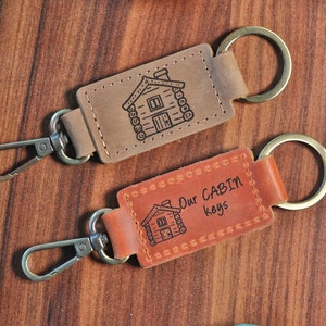 May include: Two brown leather keychains with metal key rings and a lobster clasp. One keychain has a cabin illustration and the text "Our Cabin Keys".