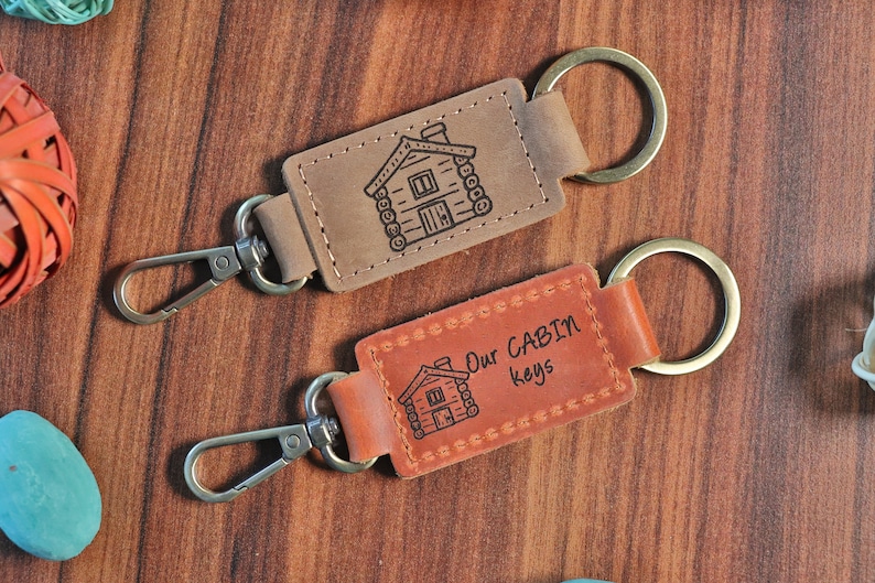 May include: Two brown leather keychains with metal key rings and a lobster clasp. One keychain has a cabin illustration and the text "Our Cabin Keys".