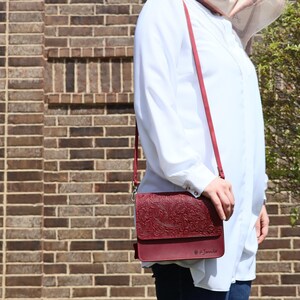 May include: A burgundy leather crossbody bag with an embossed floral design. The bag has a long strap and a flap closure. The bag is worn by a person wearing a white shirt.
