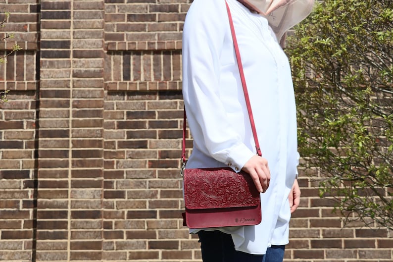 May include: A burgundy leather crossbody bag with an embossed floral design. The bag has a long strap and a flap closure. The bag is worn by a person wearing a white shirt.