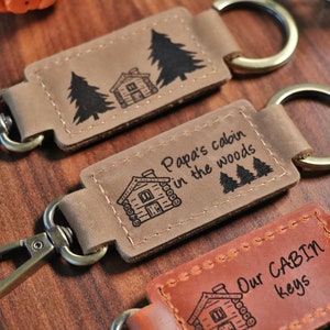 May include: Three leather keychains with cabin-themed designs. The first keychain features a cabin and two pine trees. The second keychain features a cabin and the text "Papa's cabin in the woods". The third keychain features a cabin and the text "Our CABIN keys".