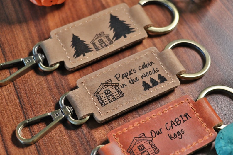 May include: Three leather keychains with cabin-themed designs. The first keychain features a cabin and two pine trees. The second keychain features a cabin and the text "Papa's cabin in the woods". The third keychain features a cabin and the text "Our CABIN keys".