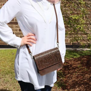 May include: A brown leather crossbody bag with an embossed floral design. The bag has a flap closure and a long adjustable strap.