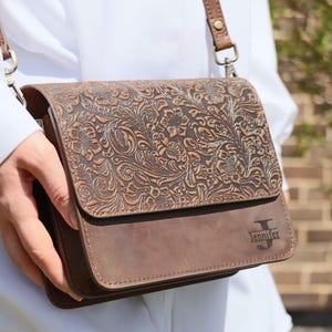 May include: A brown leather crossbody bag with an embossed floral design. The bag has a flap closure and a personalized inscription that reads "Jennifer".