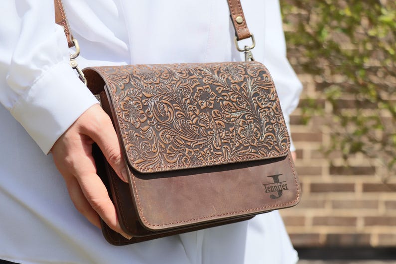 May include: A brown leather crossbody bag with an embossed floral design. The bag has a flap closure and a personalized inscription that reads "Jennifer".
