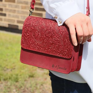 May include: A red leather crossbody bag with a floral embossed design. The bag has a flap closure and a personalized inscription that reads "Jennifer".