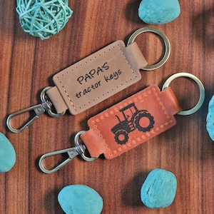 Puede incluir: Dos llaveros de cuero marrón con anillos de metal. Un llavero tiene el texto "PAPA'S tractor keys" grabado. El otro llavero tiene una silueta de un tractor grabada.