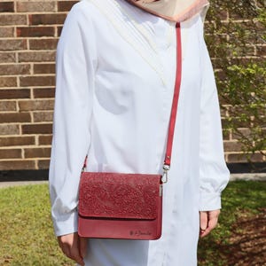 May include: A burgundy leather crossbody bag with an embossed floral pattern. The bag has a long strap and a silver clasp. The bag is worn by a person wearing a white shirt.