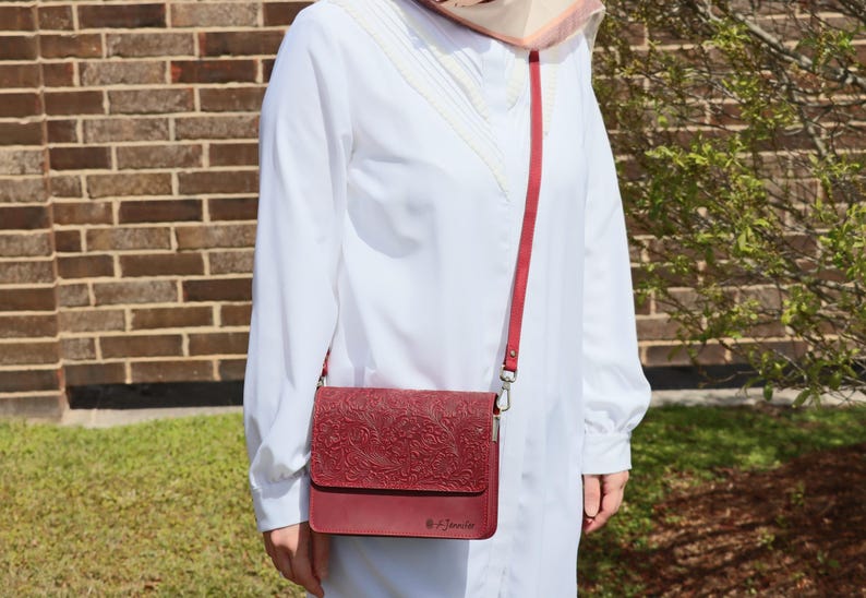 May include: A burgundy leather crossbody bag with an embossed floral pattern. The bag has a long strap and a silver clasp. The bag is worn by a person wearing a white shirt.