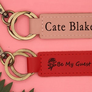 May include: Two leather keychains with gold key rings. The top keychain is pink and says "Cate Blake". The bottom keychain is red and says "Be My Guest" with a small rose design.