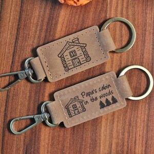 May include: Two brown leather keychains with metal rings. The top keychain has a small cabin illustration. The bottom keychain has a cabin illustration and the text "Papa's cabin in the woods".