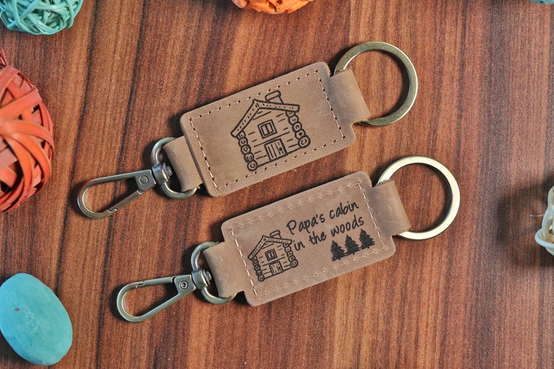 May include: Two brown leather keychains with metal rings. The top keychain has a small cabin illustration. The bottom keychain has a cabin illustration and the text "Papa's cabin in the woods".