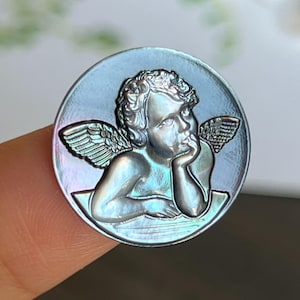 以下が含まれることがあります： 翼のある天使の像が刻まれた、虹色の青と緑の真珠貝製の丸いカメオです。天使はあごを手に乗せています。