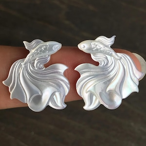 Puede incluir: Dos pendientes iridiscentes con forma de pez. Los pendientes están tallados en un material nacarado, con aletas y colas detalladas. Los peces se enfrentan, creando un diseño simétrico. Fondo neutro.