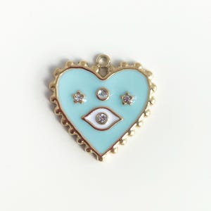 Könnte beinhalten: Ein herzförmiger Anhänger mit hellblauem Emaille-Finish und einem goldfarbenen, gewellten Rand. Der Anhänger hat ein weißes Augendesign und drei klare Kristallakzente. Oben befindet sich eine kleine Schlaufe zum Befestigen.