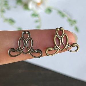 May include: Two antique gold-toned metal decorative embellishments with a delicate, intricate design. The embellishments are shaped like a stylised heart with a swirling pattern.