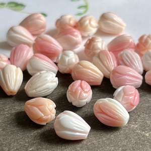 Puede incluir: Una colección de cuentas de concha de color rosa y blanco con forma de flores. Las cuentas están talladas con intrincados detalles y tienen un aspecto natural y orgánico.