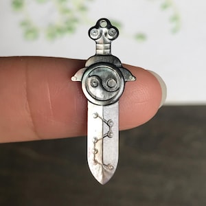 May include: A silver-toned pendant in the shape of a sword. The pendant features a Yin Yang symbol and a decorative design on the blade. The top has a loop for a chain. The pendant is held by a finger.