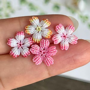 Peut inclure: Quatre petites fleurs en tissu faites à la main dans des tons de rose, blanc et orange. Les fleurs sont réalisées avec une technique délicate de nœuds et ont un aspect légèrement texturé.