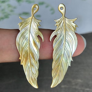 May include: Two iridescent feather-shaped pendants. The pendants have a pearlescent sheen with gold and white tones. Each feather has a loop at the top for hanging and detailed veining. The overall theme is jewelry.