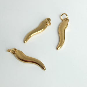 Puede incluir: Tres colgantes dorados con forma de cuerno italiano, también conocidos como cornicello, se muestran sobre un fondo blanco. Cada colgante tiene forma curva de cuerno con un pequeño lazo en la parte superior para sujetarlo a un collar o pulsera. Los colgantes son brillantes y reflejan la luz.
