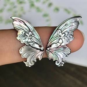 Puede incluir: Dos alas de mariposa iridiscentes con intrincados diseños florales y arremolinados. Las alas son plateadas, blancas y tienen toques de verde y morado. Ideales para joyería o proyectos de manualidades.