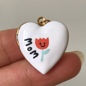May include: A heart-shaped pendant with a white enamel finish. The pendant features a red tulip with a smiling face and the word "Mom" in black. The pendant has a gold-colored border and a small loop for a chain.