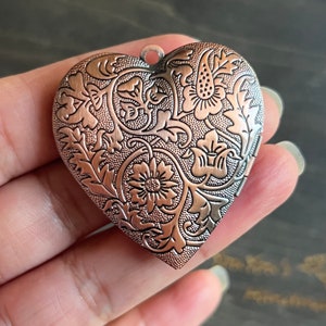 Puede incluir: Un medallón en forma de corazón con un diseño floral detallado en tonos cobre y plata. El medallón tiene una abertura con bisagra y un delicado patrón floral.
