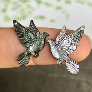 Puede incluir: Dos dijes de pájaro de color gris oscuro iridiscente con acabado de nácar. Cada pájaro se muestra en vuelo con las alas extendidas, con grabados detallados de plumas y un pequeño lazo circular en la parte superior del ala para colgar.