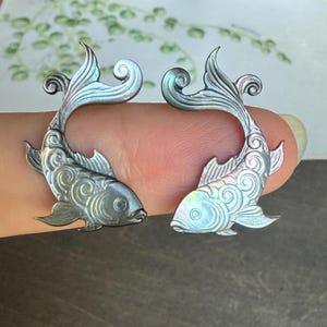May include: A pair of iridescent gray mother-of-pearl fish carvings featuring intricate swirling patterns on the scales and fins. The fish are shaped with curved tails and detailed eyes, showcasing a natural pearlescent sheen and smooth finish.