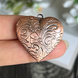 Op de afbeelding: Een koperkleurig hartvormig medaillon met een ingewikkeld bloemmotief. Het medaillon heeft een scharnierende opening en een lus om een ​​ketting aan te bevestigen.