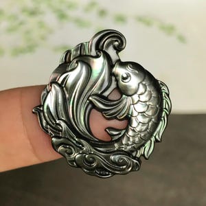 May include: A circular pendant carved from dark, iridescent shell featuring a detailed koi fish swimming in a curved motion. The surface displays a metallic sheen with hints of green and silver, showcasing intricate scale and fin textures.