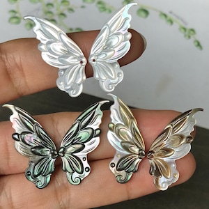 Peut inclure: Trois paires d'éléments irisés en forme d'ailes de papillon. La paire supérieure est blanche, la paire du milieu est noire et verte, et la paire inférieure est dorée et blanche. Chaque aile présente des nervures détaillées et un motif floral décoratif.