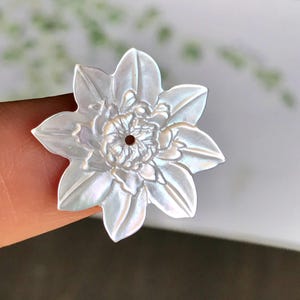 Pode incluir: Um pingente branco perolado em forma de flor com detalhes intrincados nas pétalas e no centro. O pingente tem um pequeno orifício central, sugerindo o uso na fabricação de joias ou artesanato. A flor tem uma aparência em camadas e texturizada.