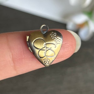 May include: A small, heart-shaped locket pendant with a brushed gold finish and a floral design. The locket has a hinged opening and is engraved with the letter "S".
