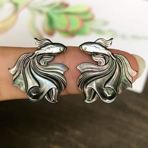 Może przedstawiać: Para srebrnych kolczyków w kształcie ryb. Każdy kolczyk ma szczegółowy wzór ryby z płynącymi płetwami i ogonem. Kolczyki mają perłowy połysk, odbijający światło.