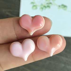 Könnte beinhalten: Drei herzförmige Verzierungen in verschiedenen Rosatönen. Jedes Herz hat ein glänzendes Finish mit einem weißen Akzent an der unteren Spitze. Die Herzen sind eng beieinander angeordnet und zeigen ihre glatten, geschwungenen Oberflächen.