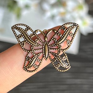 May include: A bronze-colored metal butterfly charm with intricate cut-out details. The butterfly has a delicate, filigree design and is perfect for jewelry making or crafting.