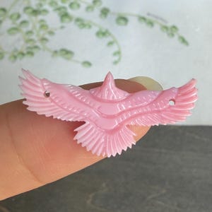 Puede incluir: Un colgante tallado de color rosa claro con forma de pájaro con las alas extendidas. El pájaro presenta tallas detalladas de plumas en sus alas y cola, e incluye dos pequeños orificios circulares, uno en cada ala, para su fijación o ensartado.
