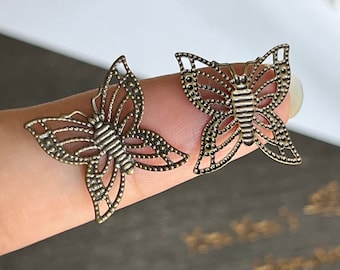 May include: Two antique gold butterfly charms with a delicate, intricate design. The charms have a cutout shape and are adorned with small, raised dots.