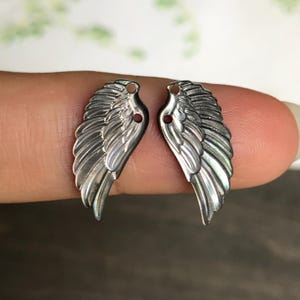 May include: Two silver-colored wing charms with detailed feather designs. Each wing has a small hole at the top for attaching to jewelry. The wings are displayed on a neutral background, showcasing their intricate craftsmanship.