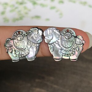 May include: Two iridescent horse-shaped pendants. Each pendant features intricate details, including a saddle with Chinese characters and a circular design. The pendants are held between fingers, showcasing their pearlescent sheen and detailed craftsmanship.