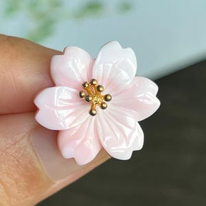 May include: A delicate pink and white cherry blossom flower charm with a gold center. The flower is carved from shell and has a glossy finish.