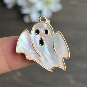 May include: A gold-toned pendant in the shape of a ghost. The ghost has a pearlescent, iridescent body with black eyes and mouth. The pendant is designed for jewelry and is suitable for Halloween.