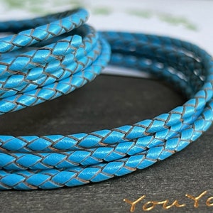Puede incluir: Primer plano de un cordón de cuero trenzado turquesa enrollado. El cordón está tejido apretadamente con un patrón visible y un borde marrón contrastante. La imagen está bien iluminada, resaltando la textura y el color del cuero. El cordón parece ser para la fabricación de joyas o manualidades.