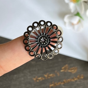 Könnte beinhalten: Ein runder, verzierter Metallschmuck mit einem kreisförmigen Design. Der Schmuck ist aus dunklem Metall mit einer goldenen Oberfläche. Er hat ein zartes, filigranes Muster aus Kreisen und Linien.